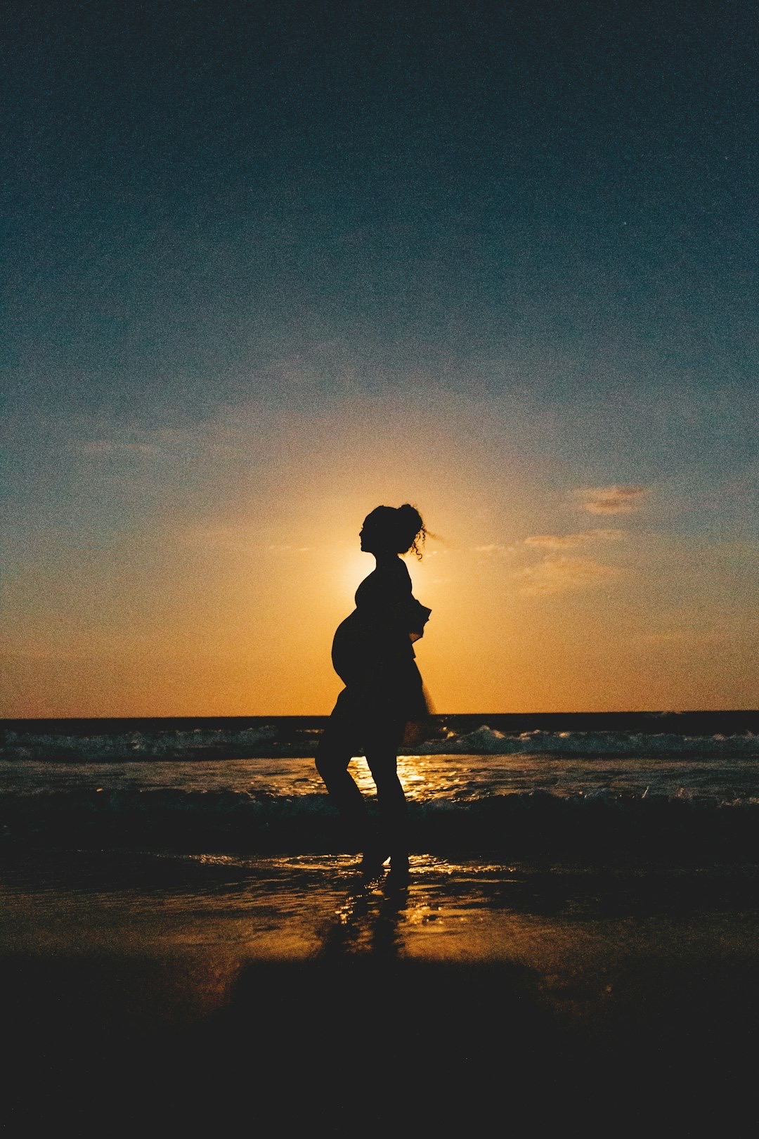 højgravid der går på stranden mens solen går ned